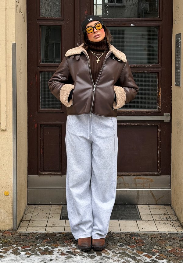 Femme portant une veste en peau retournée marron, une casquette noire, des lunettes de soleil jaunes, des colliers en couches, un col roulé marron, un pantalon gris ample, debout les mains dans les poches.