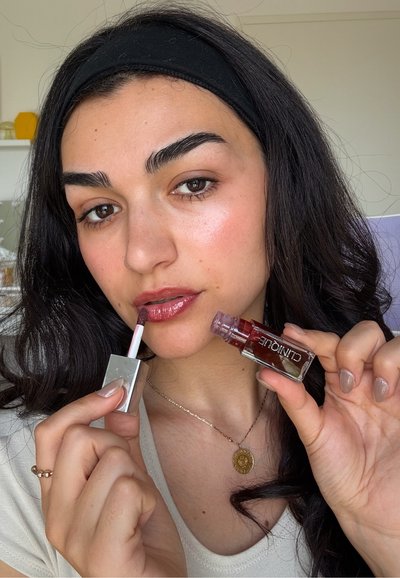 Mujer joven con cabello oscuro aplica brillo labial burdeos de Clinique, vistiendo una blusa blanca y un collar con un colgante dorado.