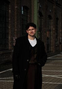 Jeune homme aux cheveux bouclés, portant des lunettes et une moustache, vêtu d'un manteau noir, de gants en cuir et de vêtements superposés, debout dans une rue urbaine.