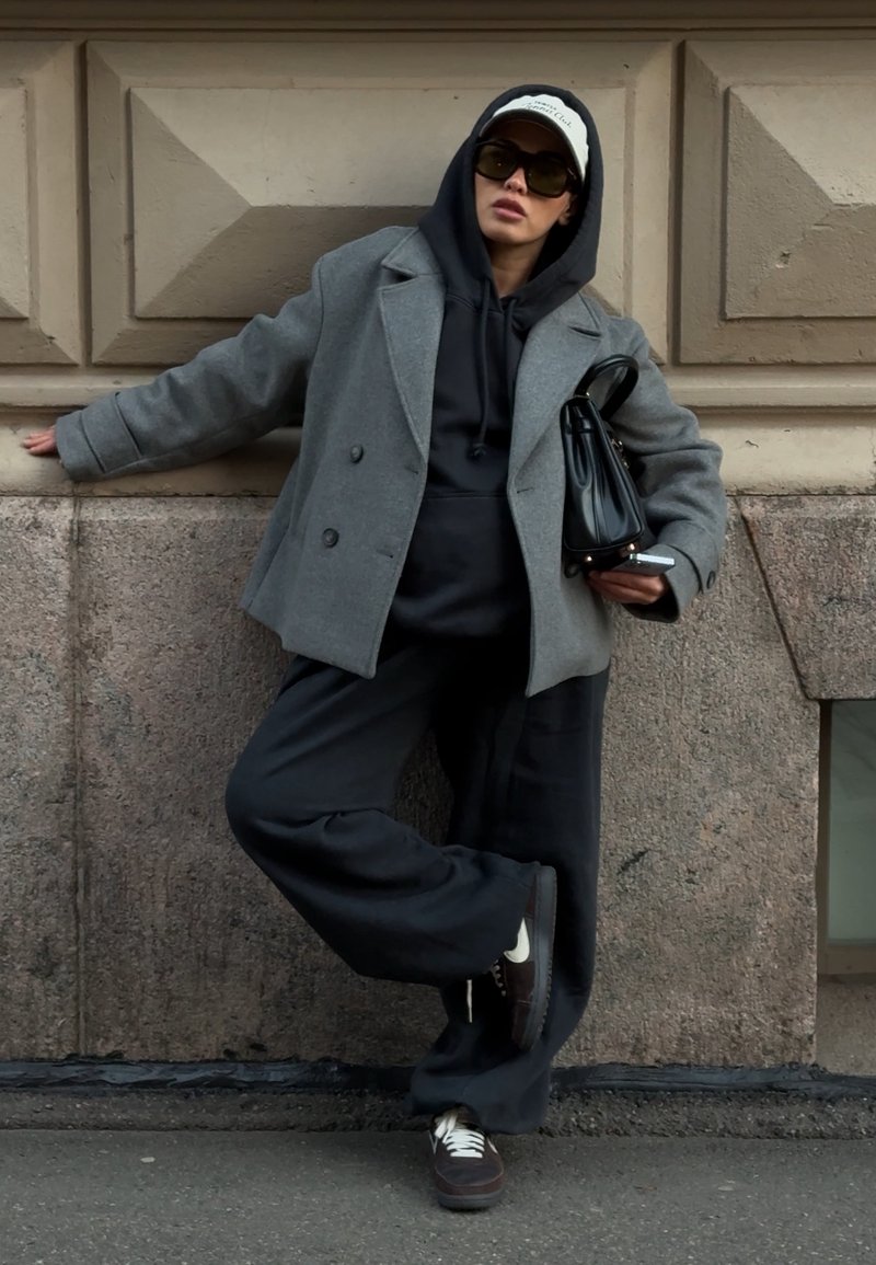 Grijze dubbelgeknoopte jas over een zwarte hoodie, gecombineerd met zwarte broek met wijde pijpen. Zwarte handtas en bruine sneakers maken de look compleet.