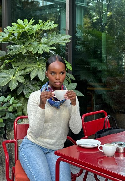 Una mujer con un suéter de punto crema y una bufanda azul con patrones sostiene una taza blanca. Ella está sentada en una mesa roja con una bolsa negra, rodeada de plantas.