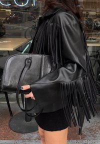 Μαύρο δερμάτινο μπουφάν με μακριά κρόσσια, συνδυασμένο με μια δομημένη μαύρη δερμάτινη τσάντα με λαβή στην κορυφή κα ι μινιμαλιστικό σχέδιο.