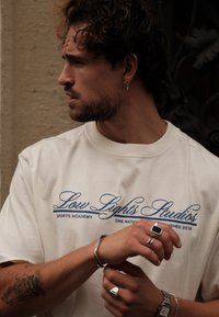 Μπεζ βαμβακερό μπλουζάκι με μπλε κείμενο "Low Lights Studios Sports Academy." Ασημί κοσμήματα στα χέρια, υποψία τατουάζ στο χέρι, υφή φόντου.