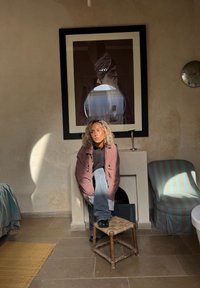 Model sidder på en halmstol iført en rød og sort ternet oversized skjorte over en sort top og blå jeans, med strukturerede vægge og møbler.