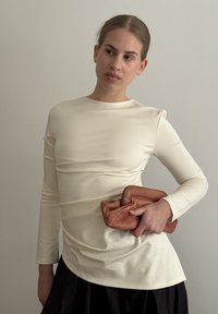 Svetlo bež dolg rokav z prilegajočo silhueto in nežno teksturo, v kombinaciji z zloženim rjavim torbico in črno krilo.