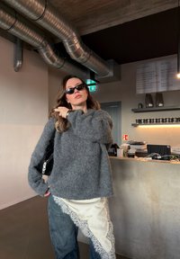 Femme portant des lunettes de soleil foncées, un pull gris en matière moelleuse et un jean avec du tissu en dentelle blanche, debout à l'intérieur d'un café moderne près d'un comptoir.