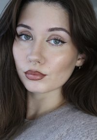 Close-up of a person with long, dark hair and light skin, wearing a gold septum ring, brown lip color, and subtle eye makeup.