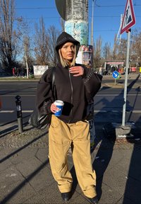 Schwarze Zip-Hoodie mit silbernen Nieten an den Schultern, beige weite Cargo-Hosen, schwarze Handtasche, hält eine Kaffeetasse. Außenbereich in urbaner Umgebung.
