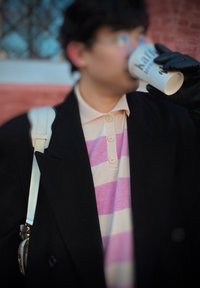 Personne en manteau noir et gants boit dans un gobelet à emporter blanc, portant une chemise rayée rose et blanche ainsi qu'un sac à bandoulière blanc.