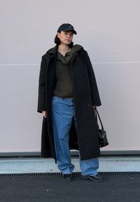 Manteau en laine noire, pull vert olive, jeans bleus amples, casquette noire et sac à main. Textures lisses, design superposé et coupe décontractée.