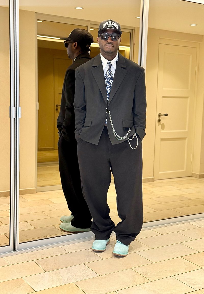 Homme portant un costume noir, une cravate à motifs, un accessoire chaîne, des chaussures turquoise, des lunettes de soleil et une casquette noire, debout près de portes coulissantes en miroir à l'intérieur.