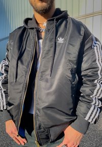 Homme portant une veste Adidas noire avec des rayures blanches sur les manches, superposée à un sweat à capuche noir et une chemise blanche, debout en plein air.