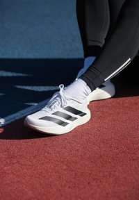 Person iført sorte leggings, hvide sokker og hvide Adidas Adizero løbesko, siddende på en rød og blå atletikbane.