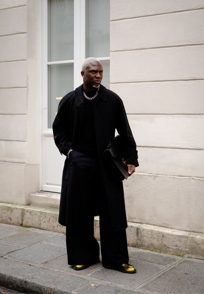 Abrigo negro, jersey negro de cuello alto, pantalones anchos negros, zapatos con detalles dorados, collar de cadena de plata, sosteniendo un bolso negro, de pie contra una pared.