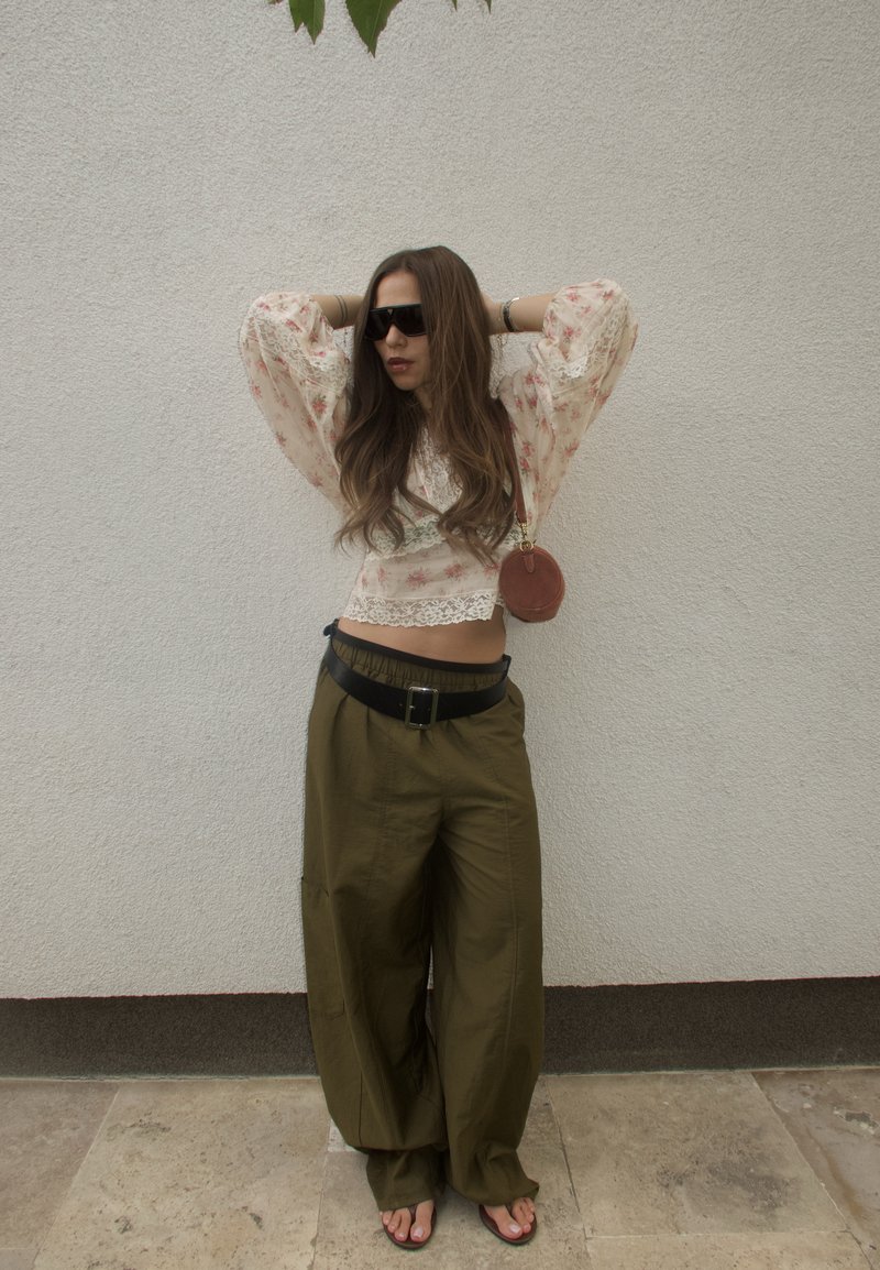 Woman wearing floral blouse, wide olive pants, black sunglasses, and sandals stands with hands behind head against plain wall.