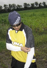 Man wearing sunglasses, a blue bandana, a yellow Puma jersey, and white arm sleeves, adjusting his sleeve in a green field with trees in the background.