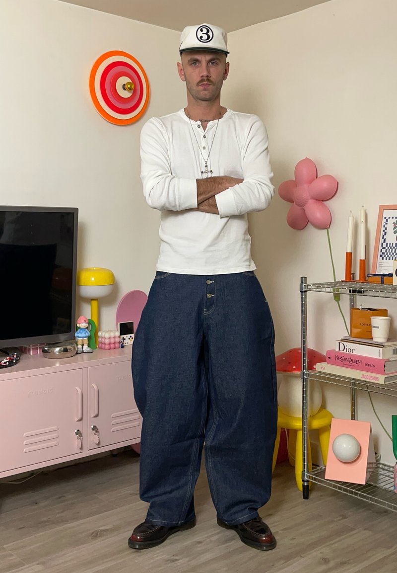 Camicia bianca a maniche lunghe, pantaloni di jeans blu larghi, scarpe nere e un berretto bianco con un numero. Sfondo con decorazioni e mobili colorati.