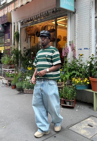 Camisa de rayas verde y blanca de manga corta, jeans sueltos de color azul claro, zapatos beige, gorra negra, de pie junto a una floristería con plantas diversas.