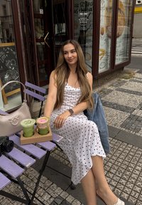 Biely šaty s bodkami, jemný materiál, dĺžka po kolená. Dvoje nápoje v priehľadných pohároch s vrstvami zelenej a ružovej farby na kartónovom nosiči. Béžová kabelka v blízkosti.