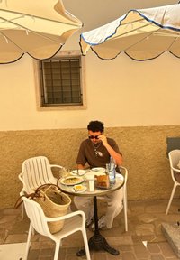 Runder Metalltisch mit einer Glasplatte und schwarzem Fuß, umgeben von weißen Plastikstühlen. Teller mit Essen, eine Wasserflasche und eine geflochtene Tasche sind zu sehen.