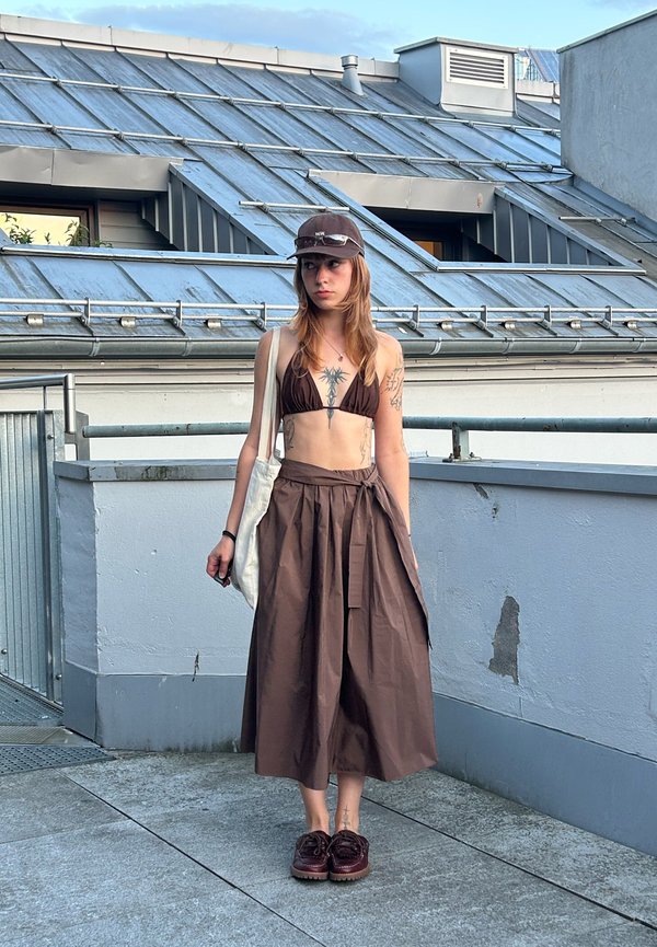 Mladá žena nosiaca hnedý bikini top, hnedú sukňu s vysokým pásom, zladené topánky a hnedú čiapku, stojaca na strešnej terase s kovovým zábradlím.