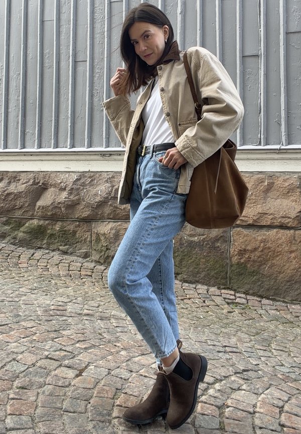 Kvinde iført beige jakke, hvid skjorte, blå jeans, mørkebrune støvler og bærende på en brun taske, står på brostensbelagt gade ved en stenmurs.