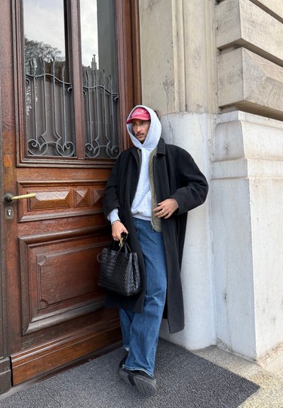 Hombre con abrigo negro, sudadera gris con capucha, gorra roja y vaqueros azules apoyado en una puerta de madera ornamentada, sosteniendo un bolso negro tejido.