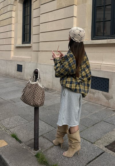 Mujer con chaqueta de cuadros, falda gris y botas beige sosteniendo una bebida, parada en la acera junto a un edificio color beige con bolso sobre un poste.