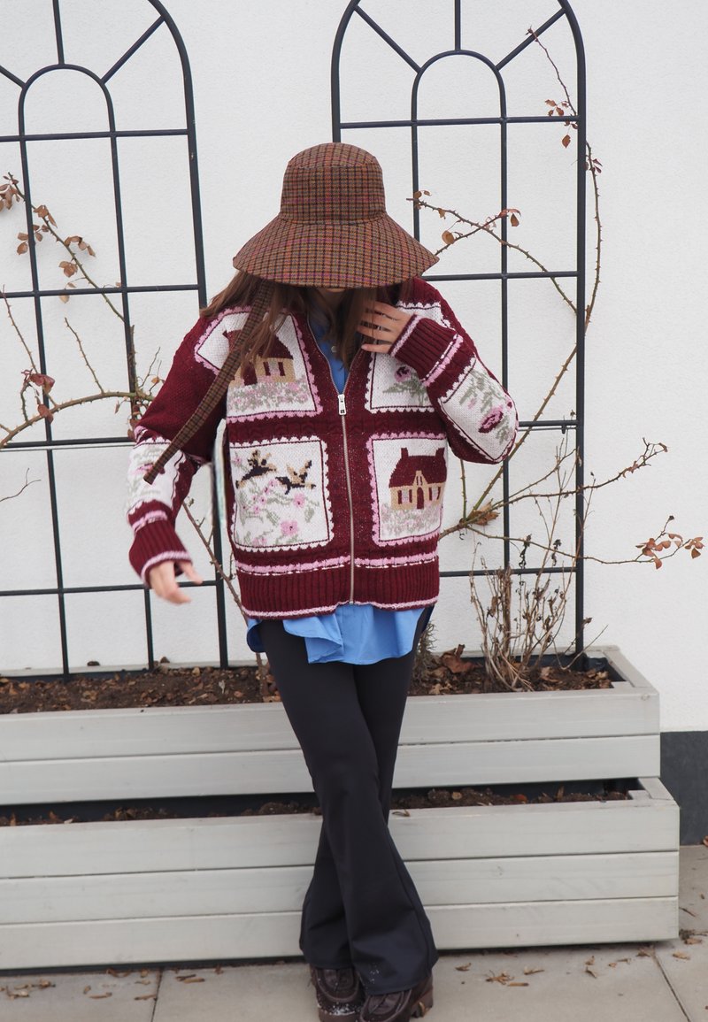 Personne portant un pull en tricot à motifs, une chemise bleue, un pantalon noir et un chapeau à larges bords à carreaux, debout près de jardinières avec des plantes grimpantes sans feuilles.