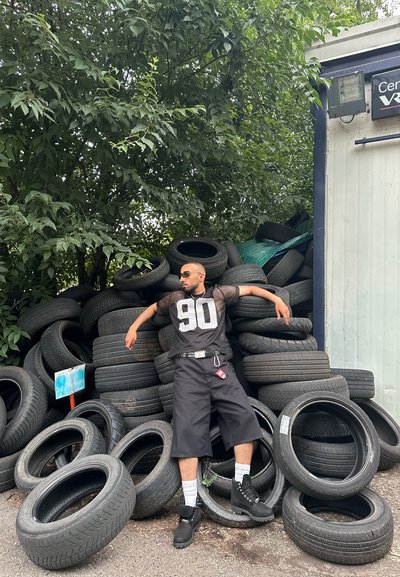 Una persona lleva puesta una camiseta de malla negra con el número "90", pantalones cortos negros y botas negras, apoyándose contra una gran pila de neumáticos desgastados.