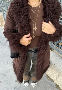 Braun pelzbesetzte Jacke über einem braunen Oberteil, kombiniert mit grauen weiten Hosen und schwarzen Sneakers, accessoriert mit geschichteten goldenen Halsketten.