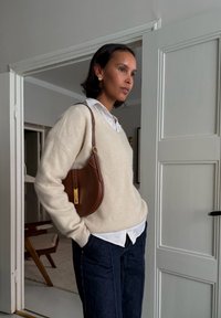 Maglione beige a maglia, camicia bianca con colletto, pantaloni in denim scuro e una borsa a tracolla in pelle marrone con un'etichetta dorata. Design semplice e contemporaneo.