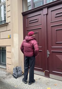 Casaco puffer cor castanho-avermelhado com capuz, calças pretas e sapatos pretos robustos; mochila cinza ao lado; de pé à frente de uma porta castanho-avermelhada.