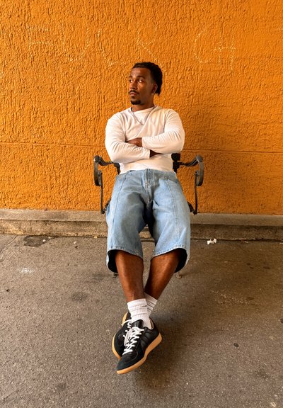 Hombre con los brazos cruzados sentado en un banco frente a una pared texturizada de color naranja brillante, vestido con una camisa blanca de manga larga, pantalones cortos de mezclilla y deportivas negras.