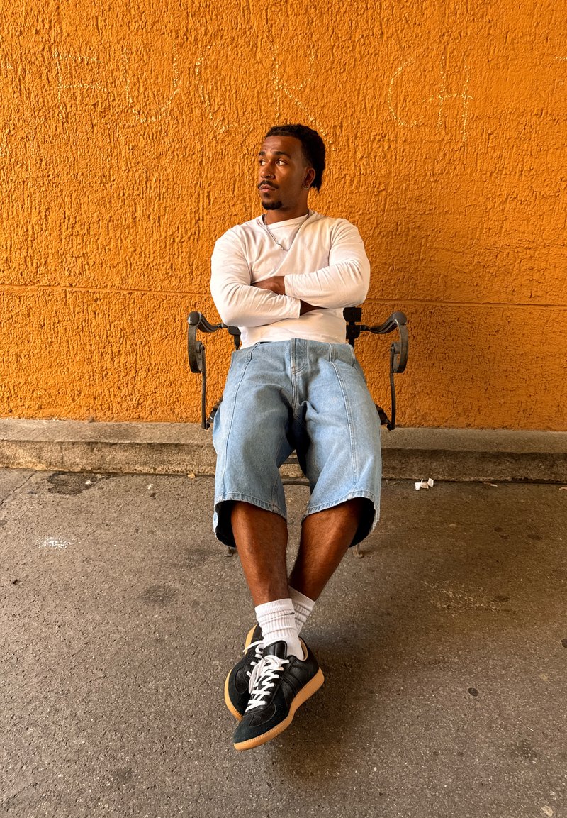Homme les bras croisés assis sur un banc contre un mur orange vif texturé, portant une chemise blanche à manches longues, un short en jean et des baskets noires.