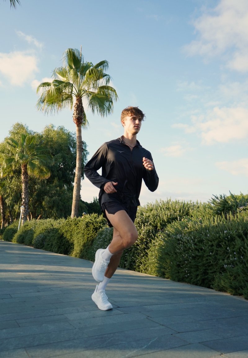 Schwarze Sportjacke, schwarze Shorts und weiße Laufschuhe. Laufen auf einem Steinweg, umgeben von grünen Sträuchern und Palmen, unter einem blauen Himmel.