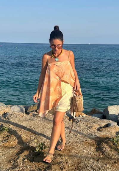 Top sin mangas de color naranja claro con estampado floral, botones y dobladillo asimétrico; shorts blancos; bolsa trenzada marrón; sandalias; fondo de mar.