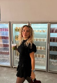 Svart kort kjole med pufferm, holder en drikke, går forbi glassfrontede kjøleskap som viser ulike hårpleieprodukter.