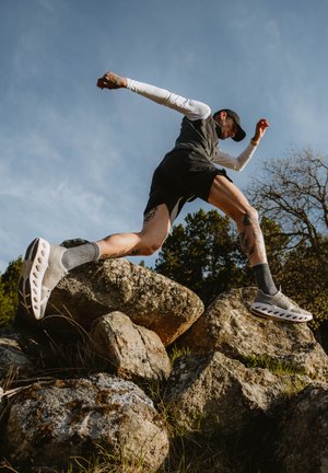 Scarpe sportive con una tomaia grigia chiaro e ritagli, suola bianca, indossate con calze grigie e pantaloni corti neri, mentre saltano su un terreno roccioso.