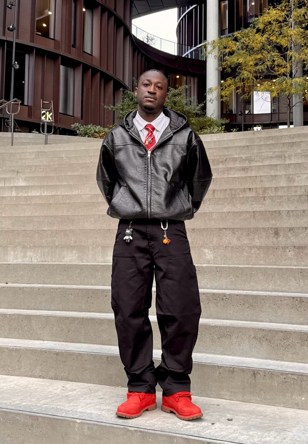 Czarna skórzana bluza z kapturem na zamek, biała koszula, czerwony krawat, czarne spodnie cargo z breloczkami oraz czerwone buty. Stoi na betonowych schodach.