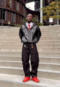 Czarna skórzana bluza z kapturem na zamek, biała koszula, czerwony krawat, czarne spodnie cargo z breloczkami oraz czerwone buty. Stoi na betonowych schodach.