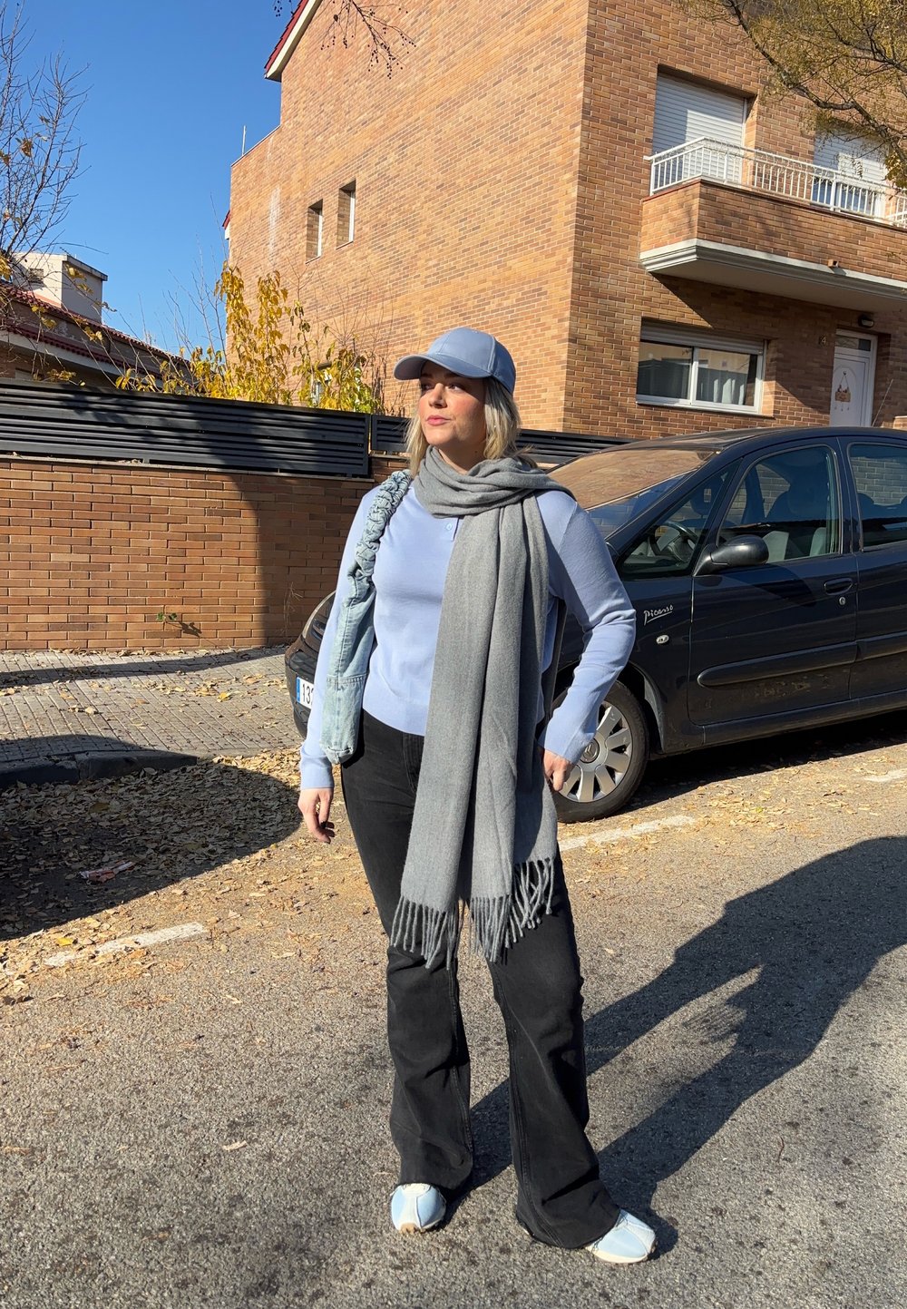 Femme portant une casquette bleu clair, un pull, une écharpe grise, un pantalon noir et des baskets blanches, debout dans une rue ensoleillée près d'une voiture noire garée et d'un bâtiment en briques.
