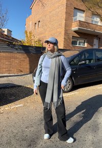 Femme portant une casquette bleu clair, un pull, une écharpe grise, un pantalon noir et des baskets blanches, debout dans une rue ensoleillée près d'une voiture noire garée et d'un bâtiment en briques.