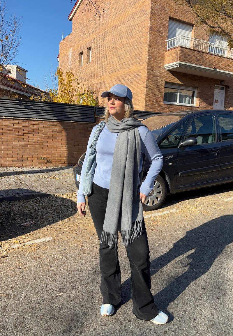 Femme portant une casquette bleu clair, un pull, une écharpe grise, un pantalon noir et des baskets blanches, debout dans une rue ensoleillée près d'une voiture noire garée et d'un bâtiment en briques.