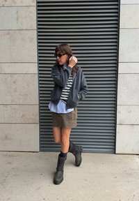 Sudadera gris con cremallera sobre una camiseta a rayas, combinada con una minifalda caqui y botas negras hasta la rodilla. Accesorios con gafas de sol.