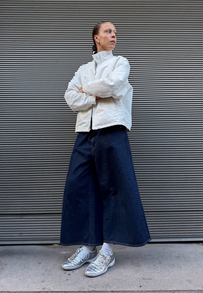 Personne les bras croisés portant une veste blanche, un jean large foncé, des chaussettes blanches et des baskets argentées, debout contre un mur rayé.