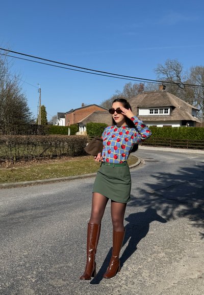 Mujer con suéter azul con manzanas rojas, falda verde, botas marrones hasta la rodilla, gafas de sol, sosteniendo un bolso marrón en una calle suburbana soleada.