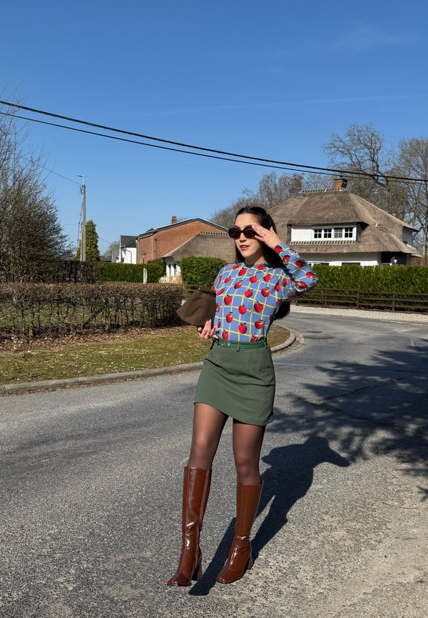 Vrouw draagt een blauwe trui met rode appels, groene rok, bruine kniehoge laarzen en een zonnebril, en houdt een bruine clutch vast op een zonnige buitenwijkstraat.