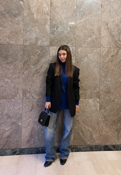 Blazer negro oversize sobre un suéter de cuello alto azul, combinado con jeans azules holgados y botas negras de tacón. Sosteniendo un bolso pequeño negro. Fondo de mármol.