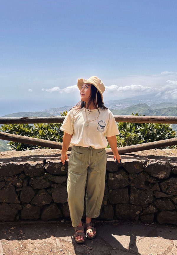 Woman wearing a bucket hat, beige t-shirt, and green pants stands by a stone wall with wooden railing, overlooking a mountainous landscape.